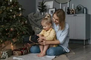 A mother and daughter joyfully open Christmas gifts by a decorated tree indoors.